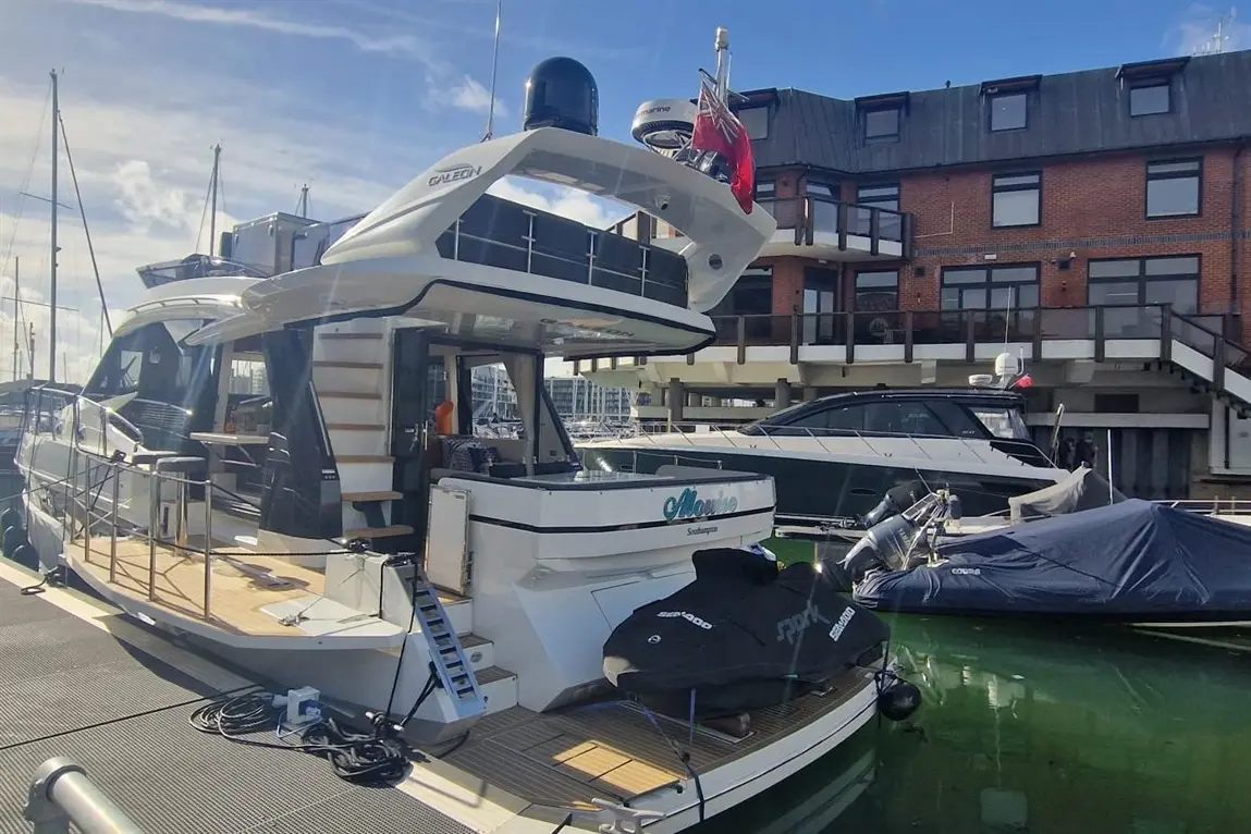 Luxury yacht docked at a marina, with a building in the background and a jet ski on the deck, under a clear blue sky.